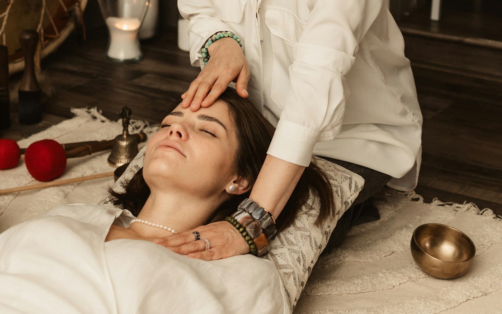 Praticienne vêtue de blanc posant délicatement les mains sur le front d'une femme allongée et détendue, bougie et bol chantant à proximité.